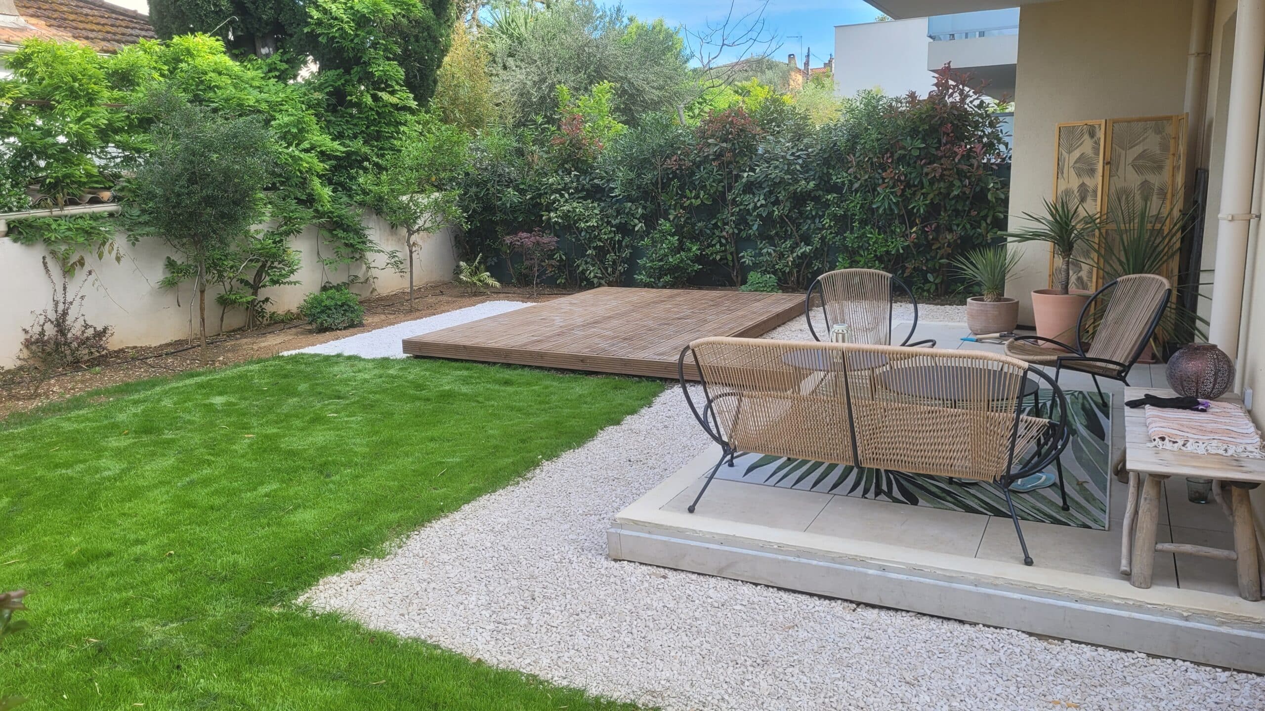 Jardin moderne avec pelouse, terrasse en bois, mobilier en rotin et gravier blanc entourés de verdure.