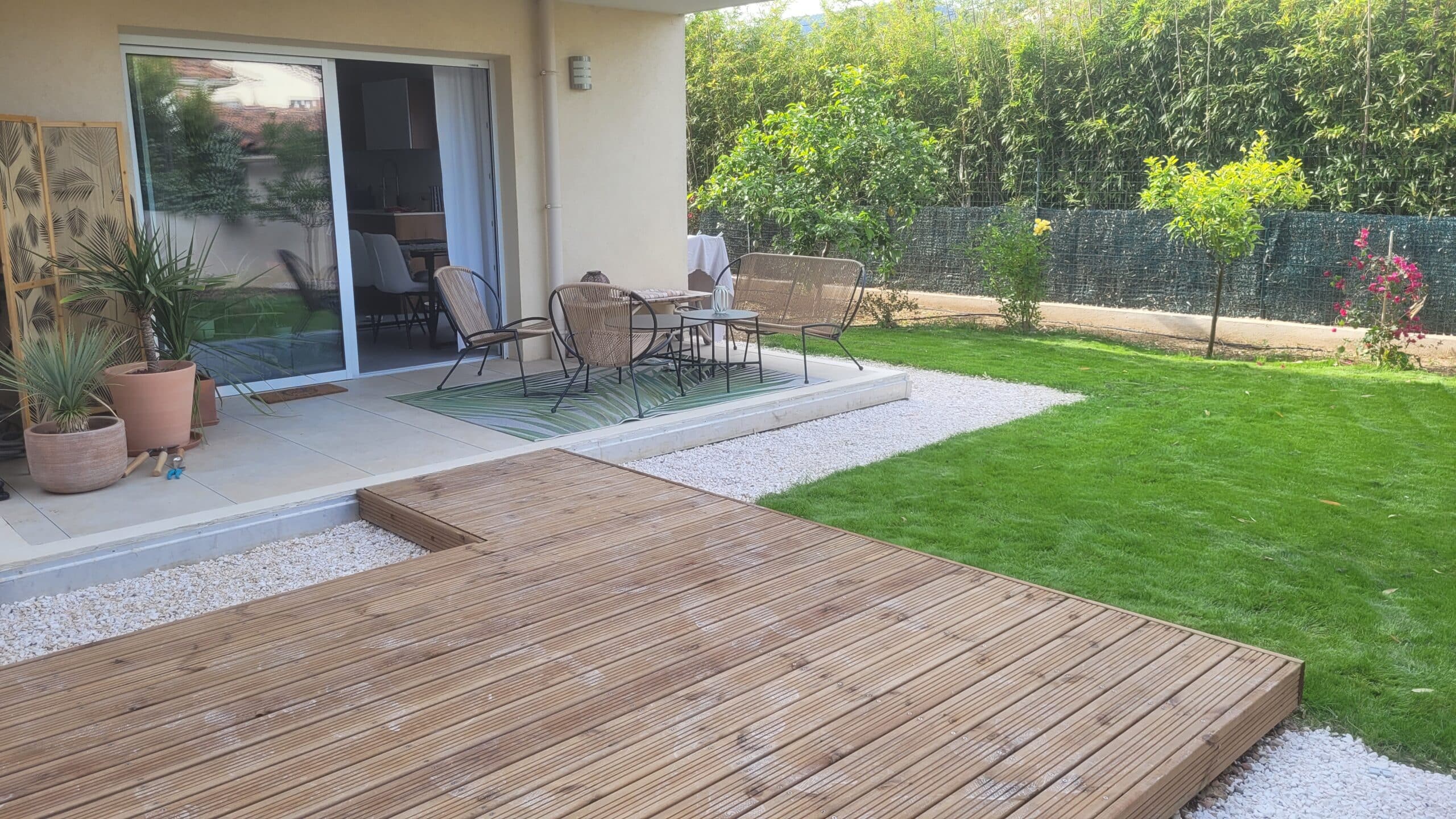 Terrasse moderne avec deck en bois, salon de jardin sur tapis et pelouse verte bordée de gravier blanc.