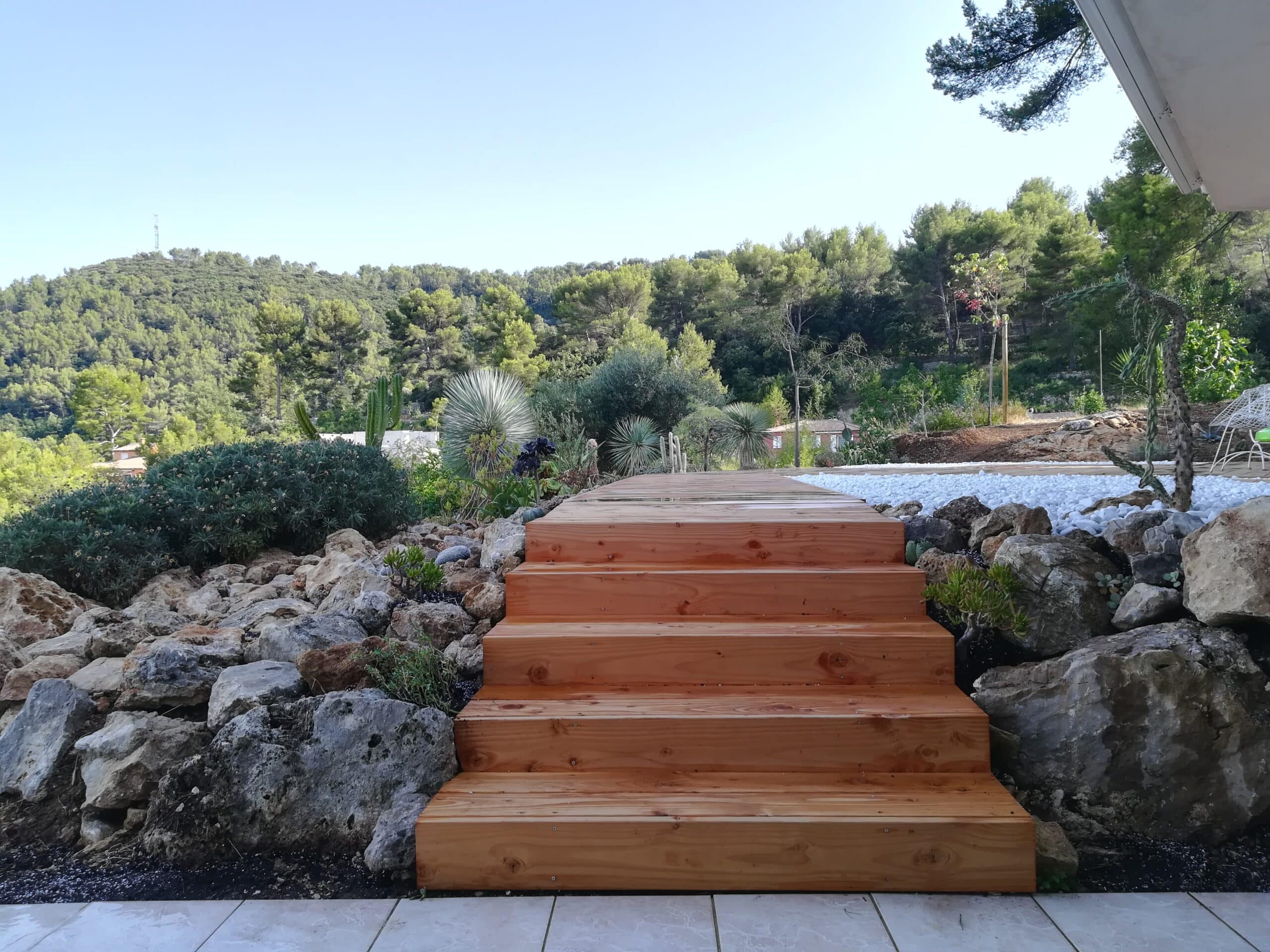 Escalier en bois clair au cœur d'un jardin sec avec rochers, succulentes et vue panoramique sur la forêt.