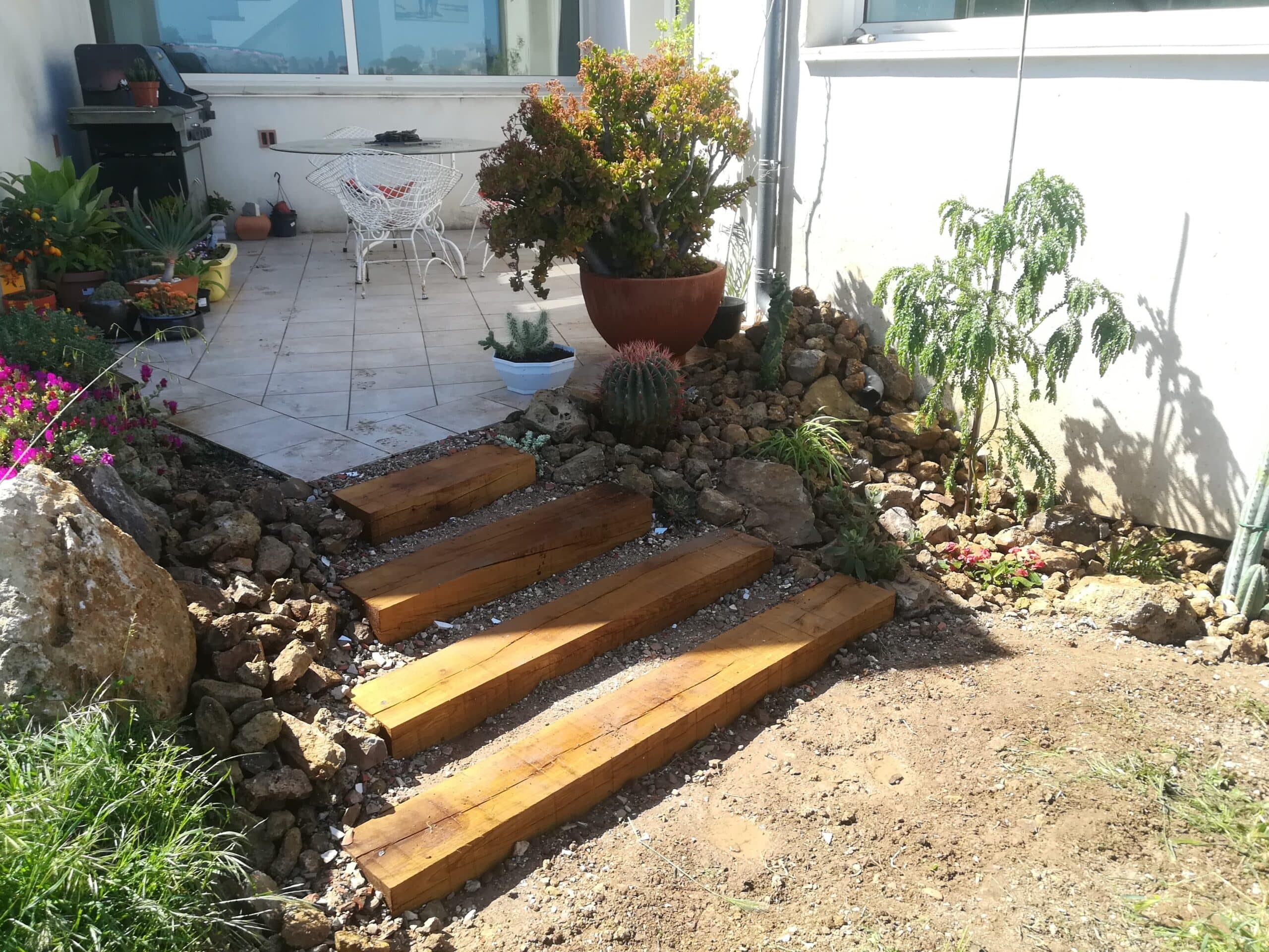 Escalier en poutres de bois, jardin de rocaille, plantes succulentes et terrasse avec mobilier blanc.