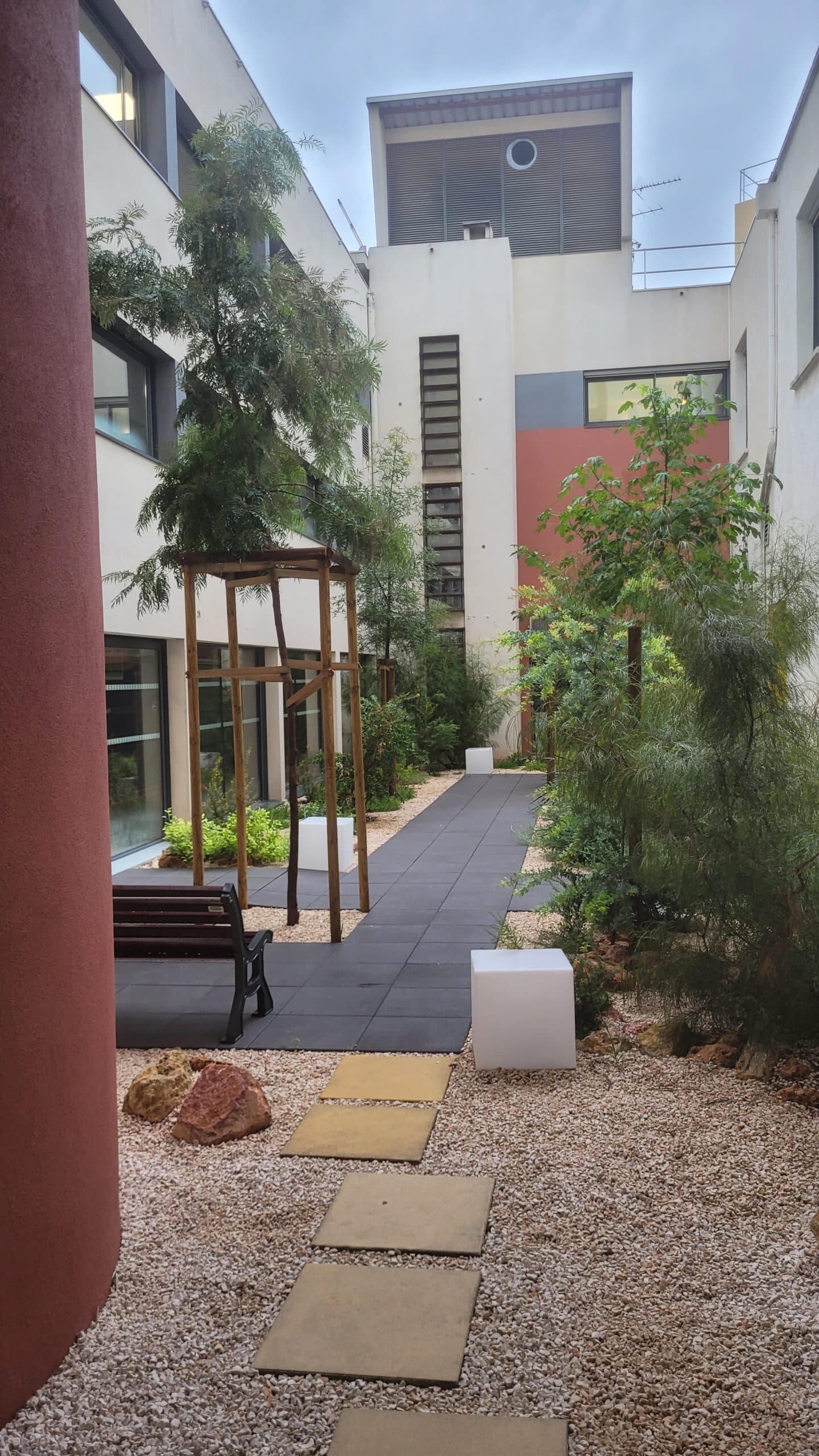 Cour intérieure moderne avec chemin pavé gris, banc en bois, cubes blancs et arbres sous un ciel nuageux.