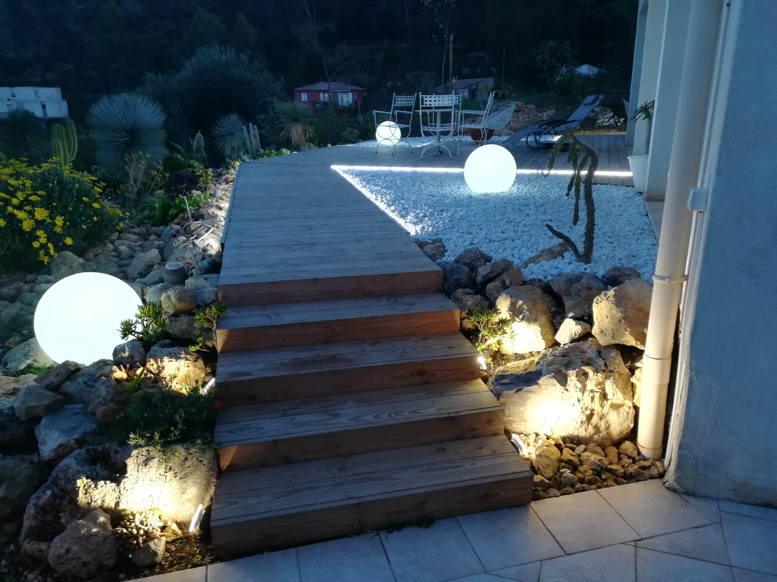 Escalier en bois et terrasse moderne avec luminaires sphériques blancs, gravier et plantes dans un jardin de nuit.