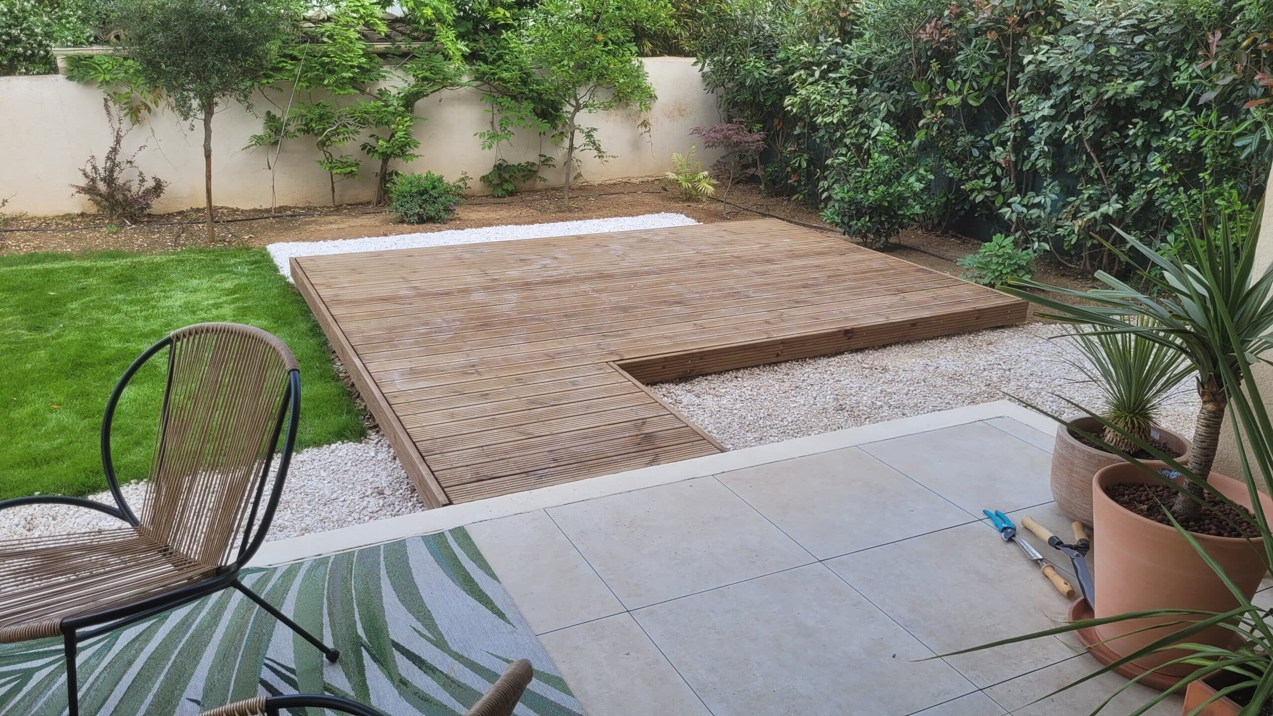 Terrasse en bois sur gravier blanc, pelouse verte et plantes en pot dans un jardin paysager moderne.