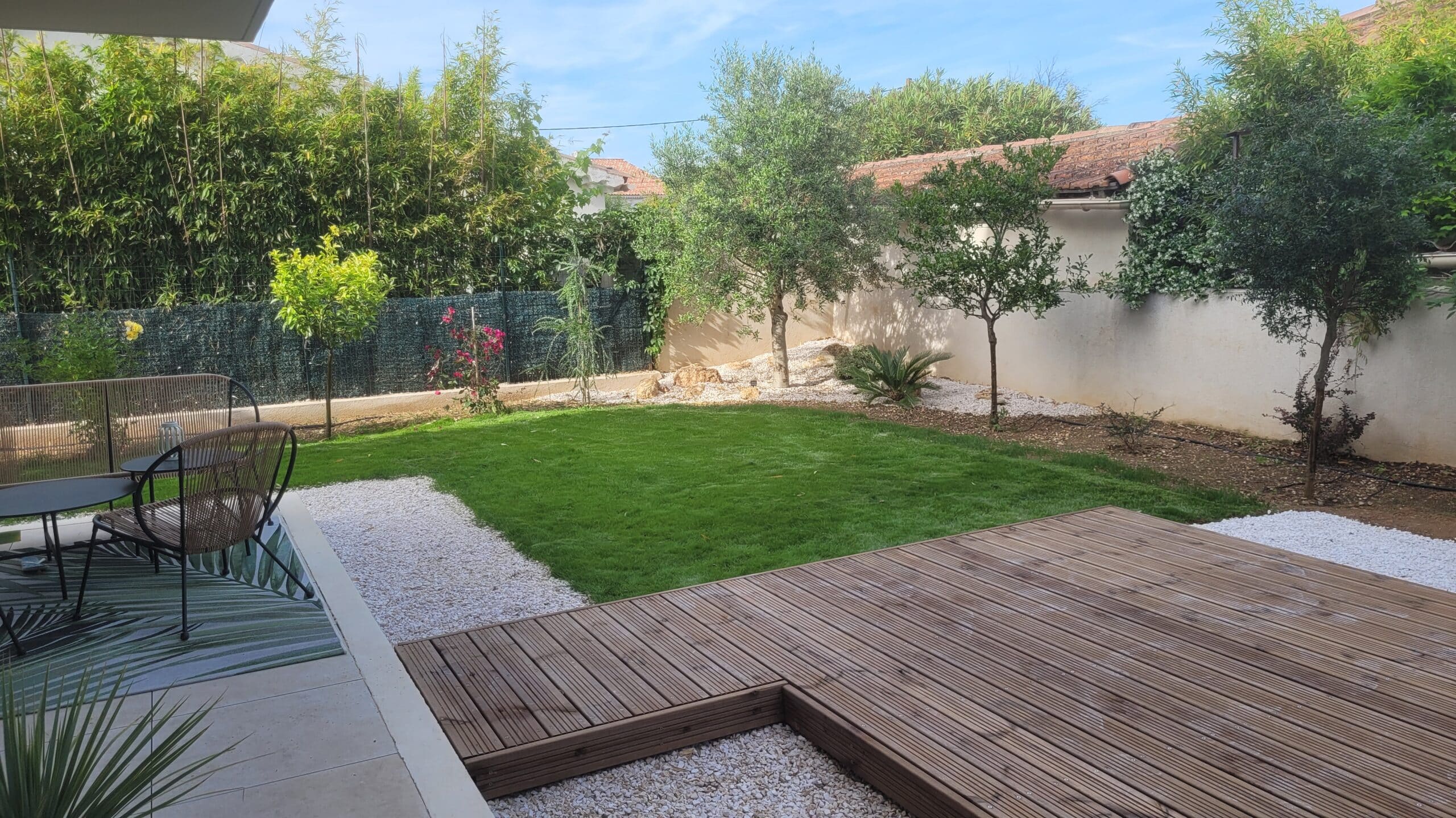 Jardin paysager avec terrasse en bois, pelouse verte, gravier blanc et mobilier de détente moderne sous le soleil.