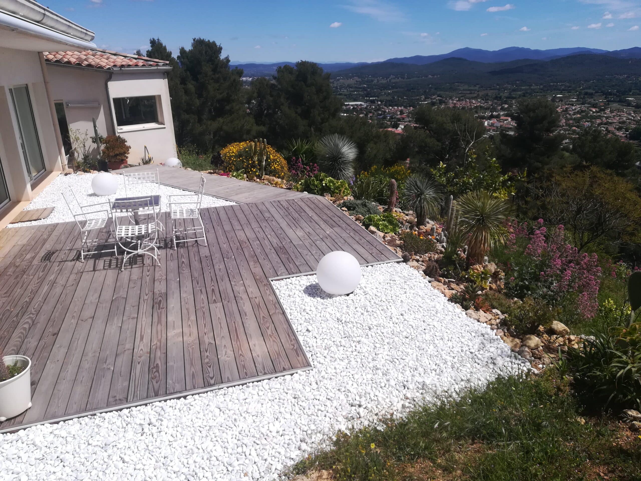 Terrasse moderne avec deck en bois, galets blancs et mobilier de jardin blanc, offrant une vue panoramique sur la vallée.