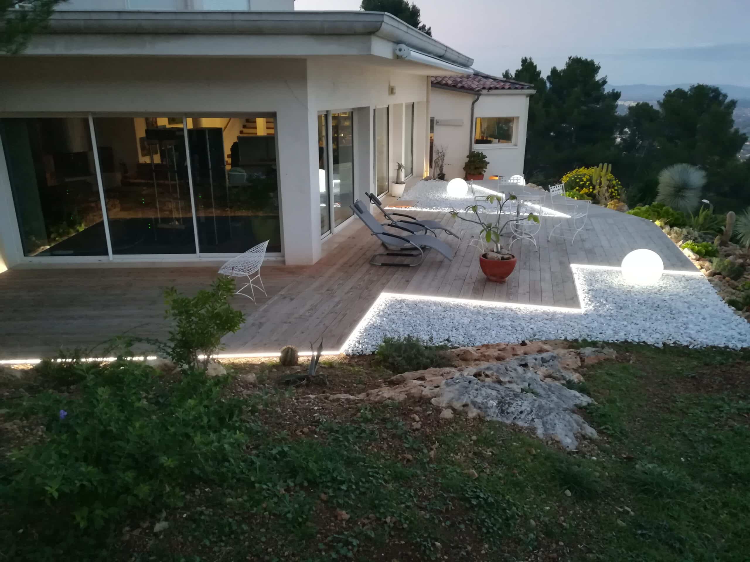 Terrasse moderne avec éclairage LED, galets blancs et chaises longues devant une maison contemporaine au crépuscule.